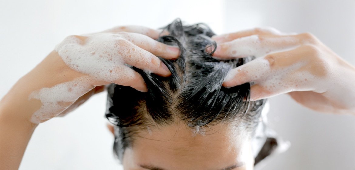 Ansicht auf den Scheitel einer dunkelhaarigen Frau. Sie shampooniert mit beiden Händen ihre Haare.